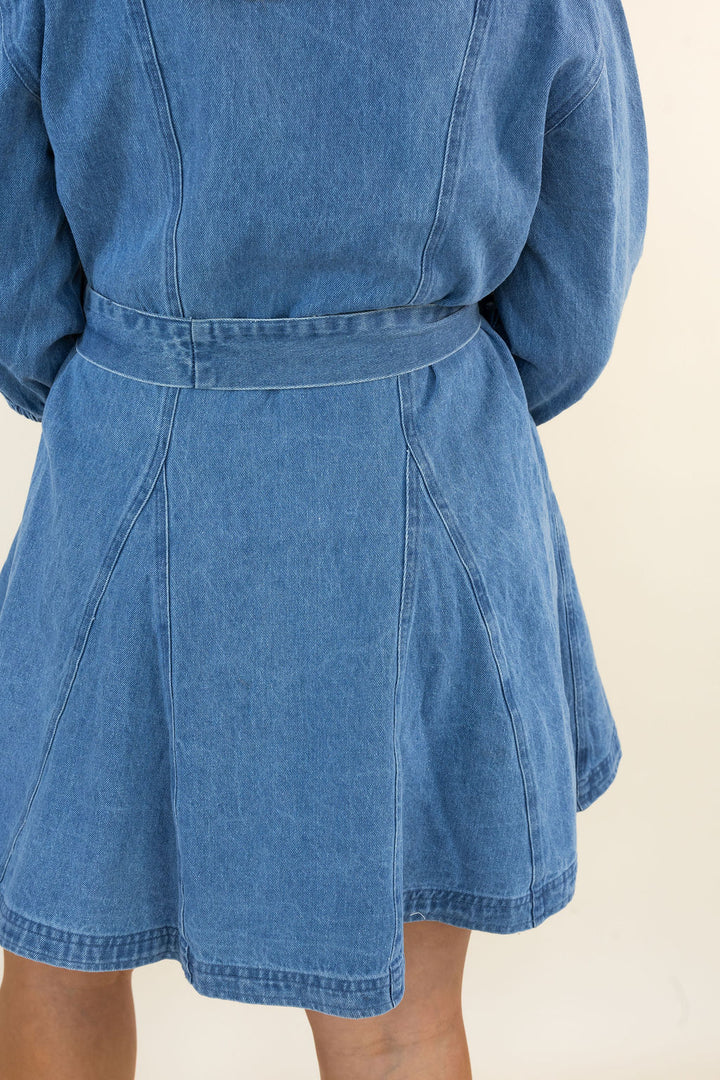 Close-up back view of a person wearing a blue denim belted dress with a flared skirt, against a plain light beige background.