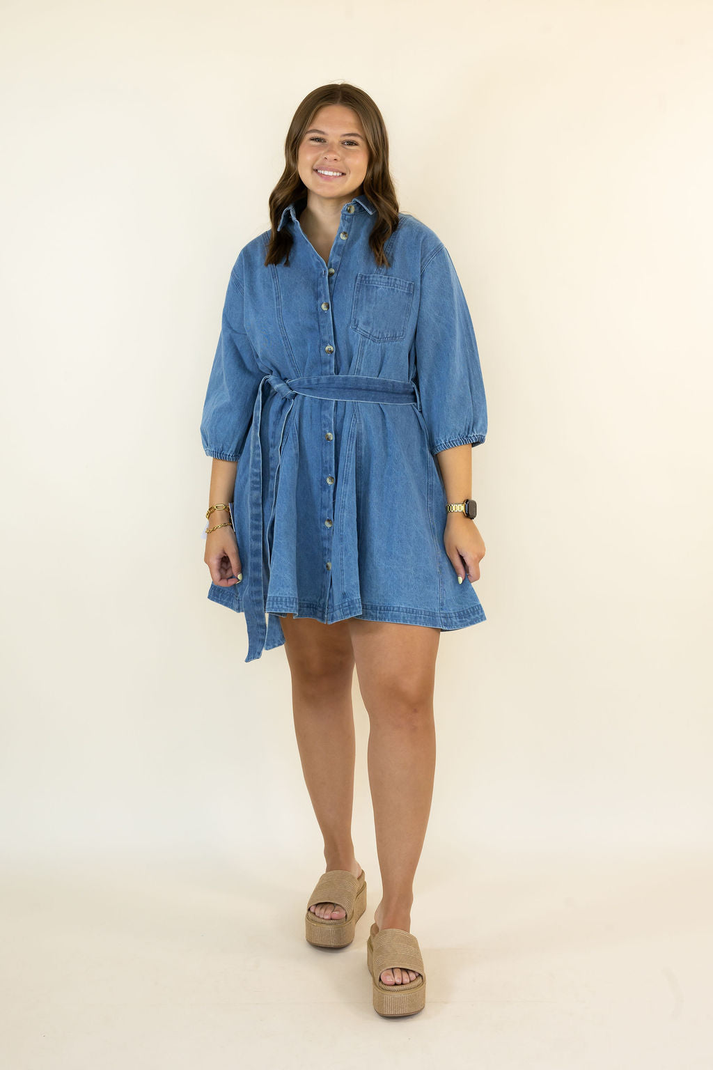 A woman modeling a blue denim shirt dress with three-quarter sleeves, a button-down front, and a self-tie waist belt, paired with beige platform sandals against a plain cream background.