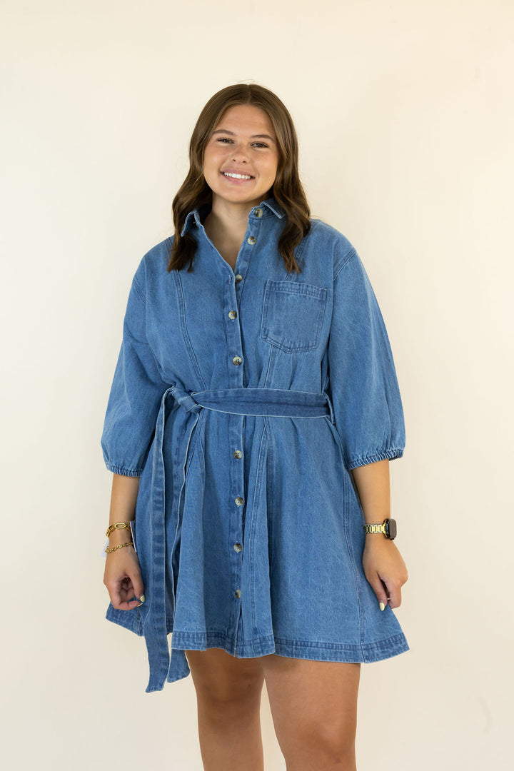A woman modeling a blue denim shirt dress with a tie waist and button front, standing against a plain off-white background.