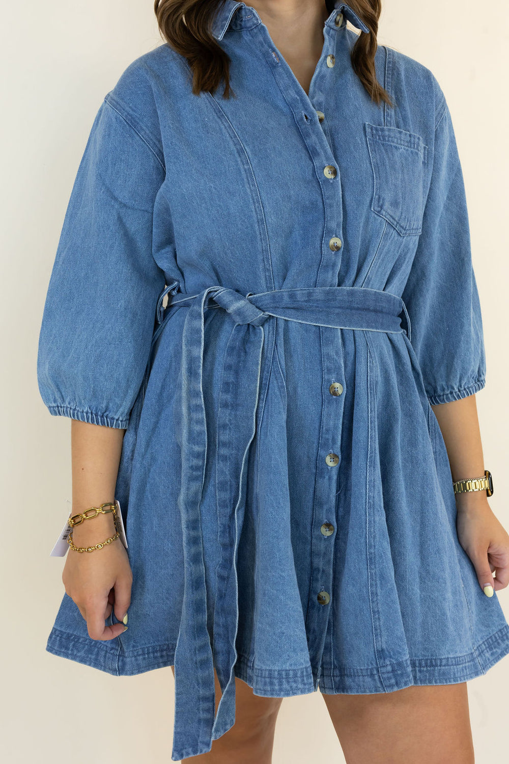 A woman wearing a blue denim shirt dress with a tie waist belt, 3/4 sleeves, and front buttons, shown against a plain light background.