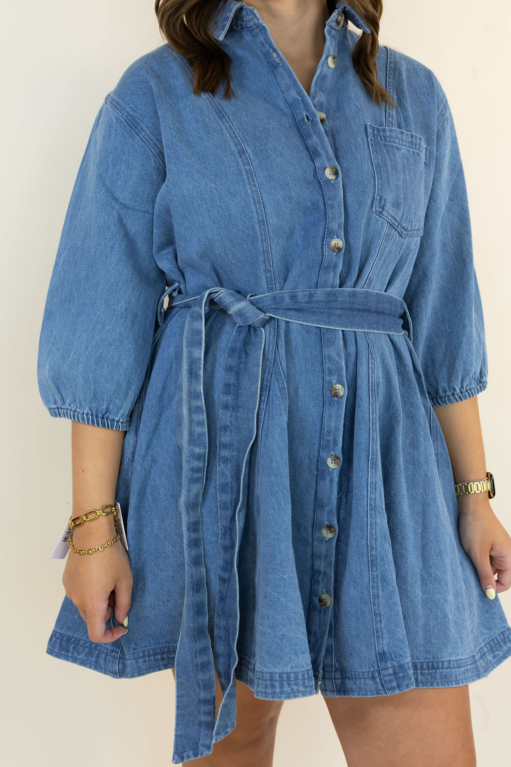 A woman wearing a blue denim shirt dress with a tie waist belt, 3/4 sleeves, and front buttons, shown against a plain light background.