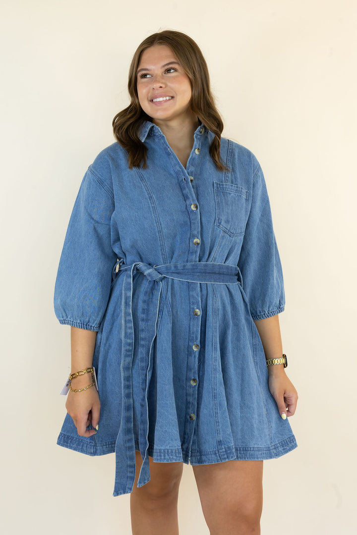 A woman modeling a blue denim shirt dress with a tie waist and button front, standing against a plain cream background.