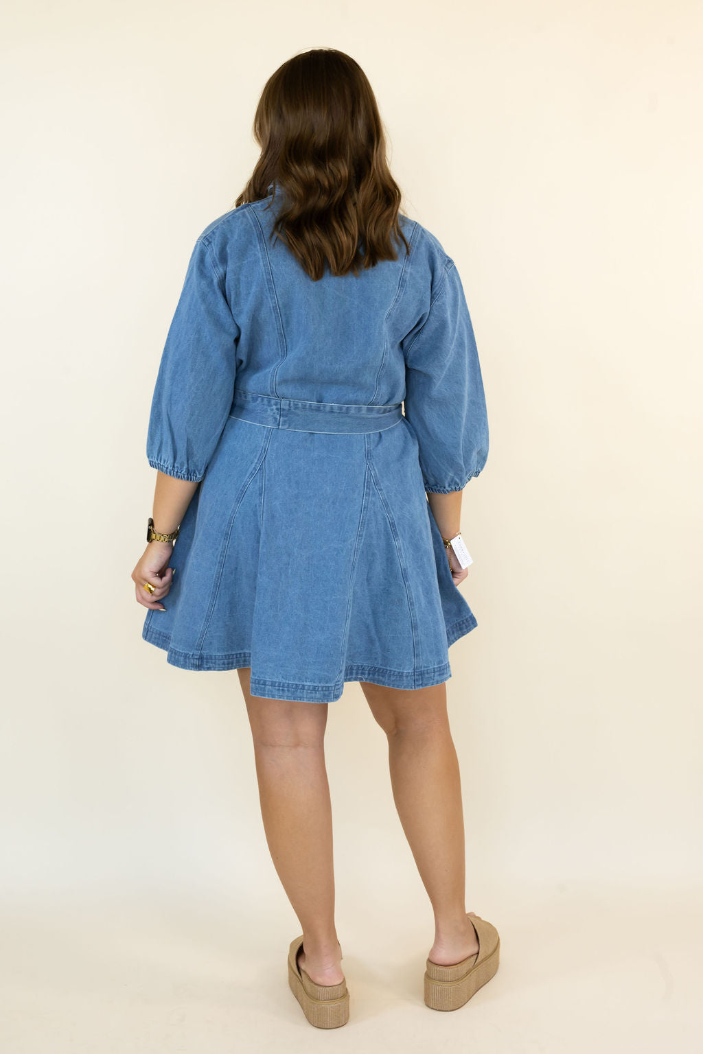 A woman viewed from behind wearing a light blue denim dress with puff sleeves and a matching belt, paired with tan platform sandals, against a plain cream background.