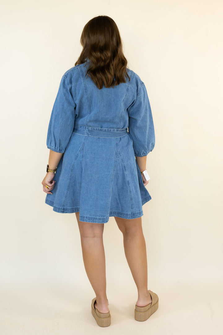 A woman viewed from behind wearing a light blue denim dress with puff sleeves and a matching belt, paired with tan platform sandals, against a plain cream background.