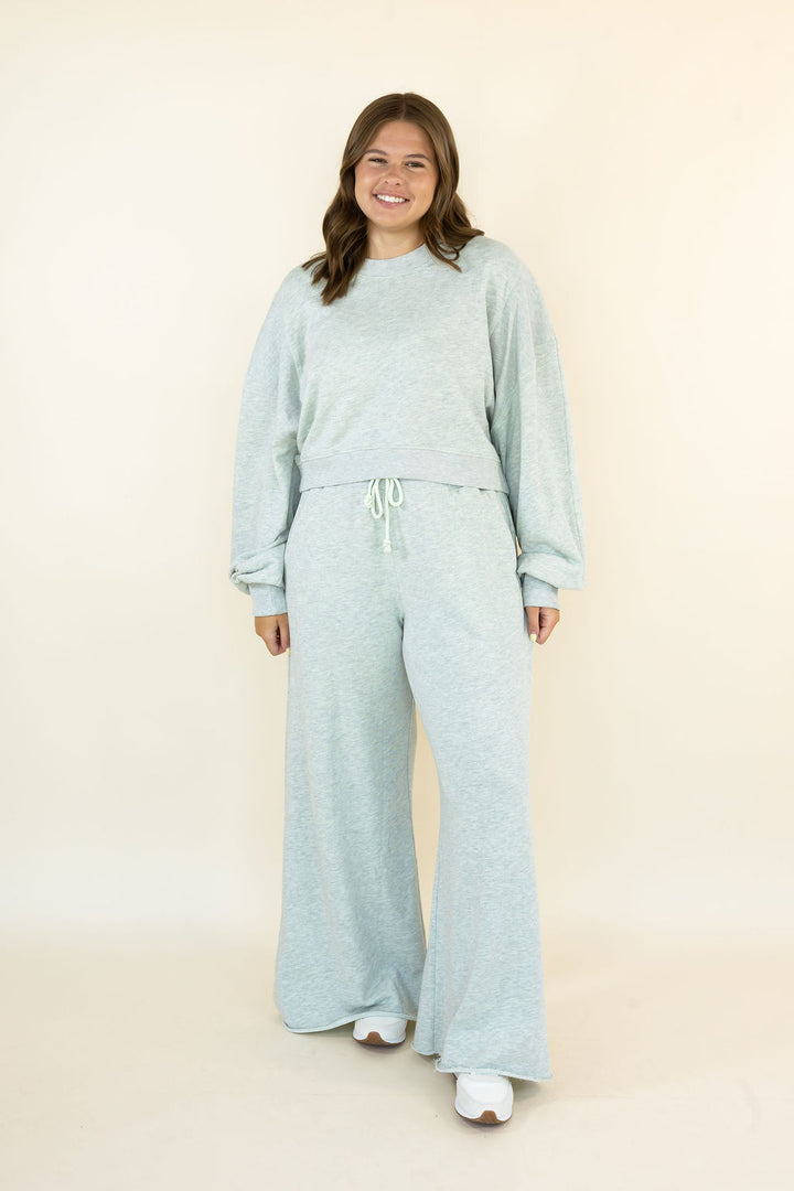 A woman modeling a light heather grey two-piece lounge set consisting of a long-sleeve cropped sweatshirt and matching wide-leg sweatpants, standing against a plain cream background.