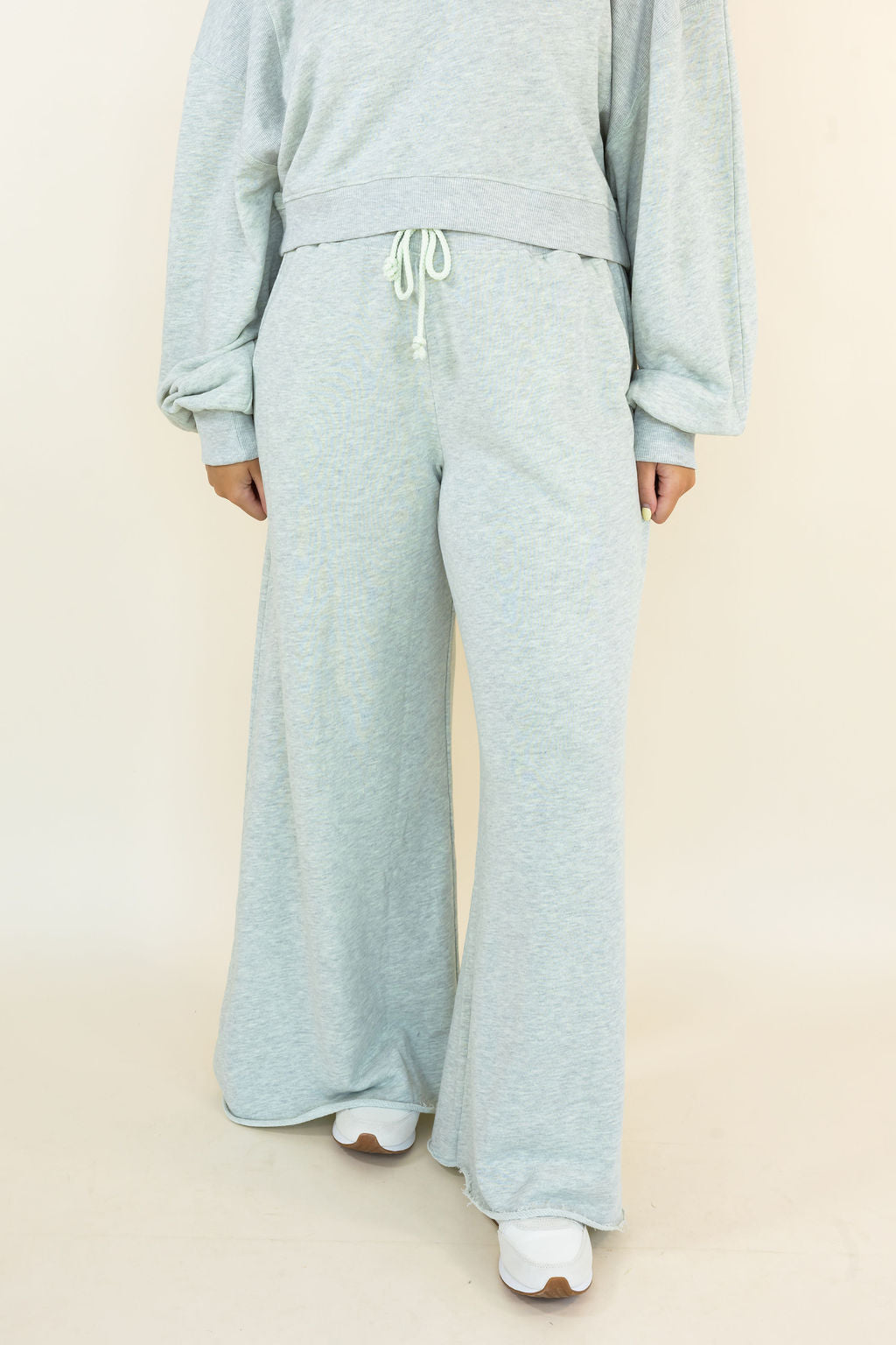A person wearing a light heather gray sweatshirt and matching wide-leg sweatpants with white sneakers, shown from the neck down against a plain beige studio background.
