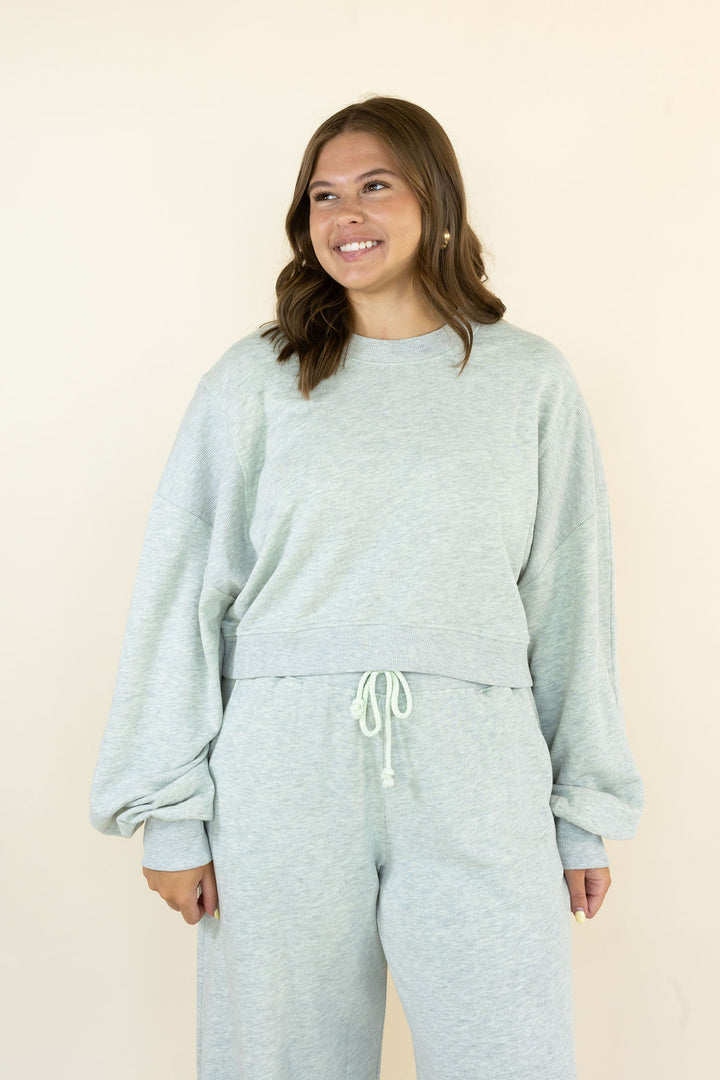 A woman modeling a light grey heathered lounge set consisting of a cropped long-sleeve sweatshirt and matching sweatpants, set against a plain beige background.