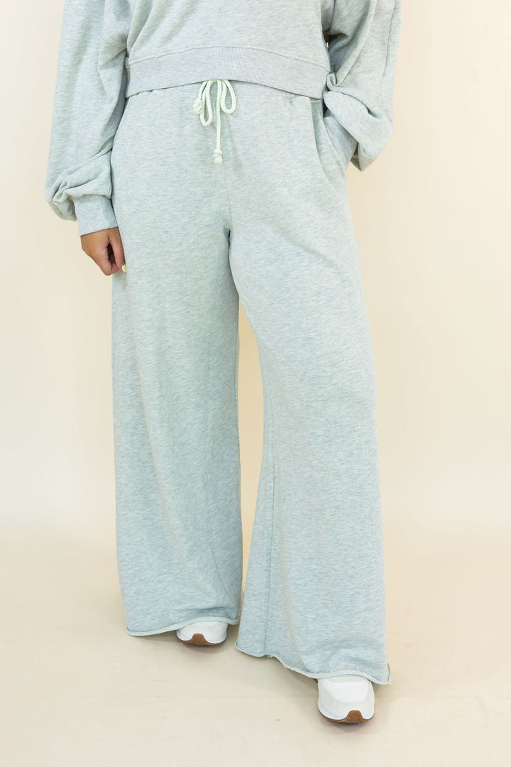 A person wearing light heather gray wide-leg sweatpants with a drawstring waist, paired with a matching gray sweatshirt and white sneakers, against a plain beige studio background.