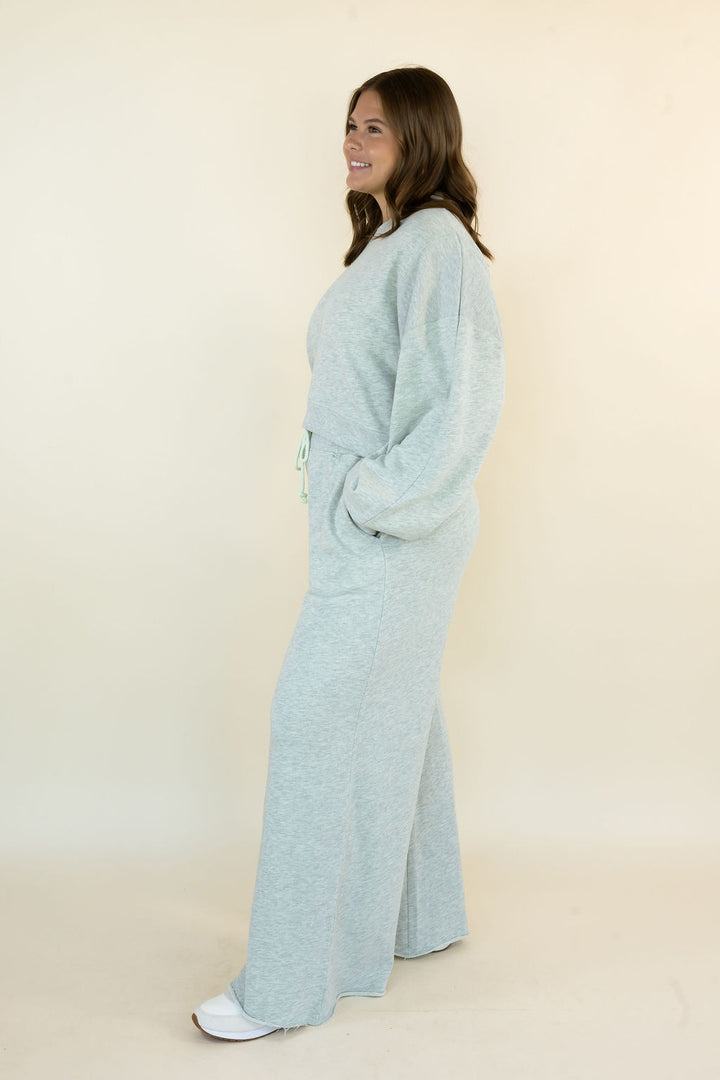 A woman wearing a light heather grey two-piece lounge set consisting of a long-sleeve crewneck top and matching wide-leg pants, standing in profile against a plain cream background.