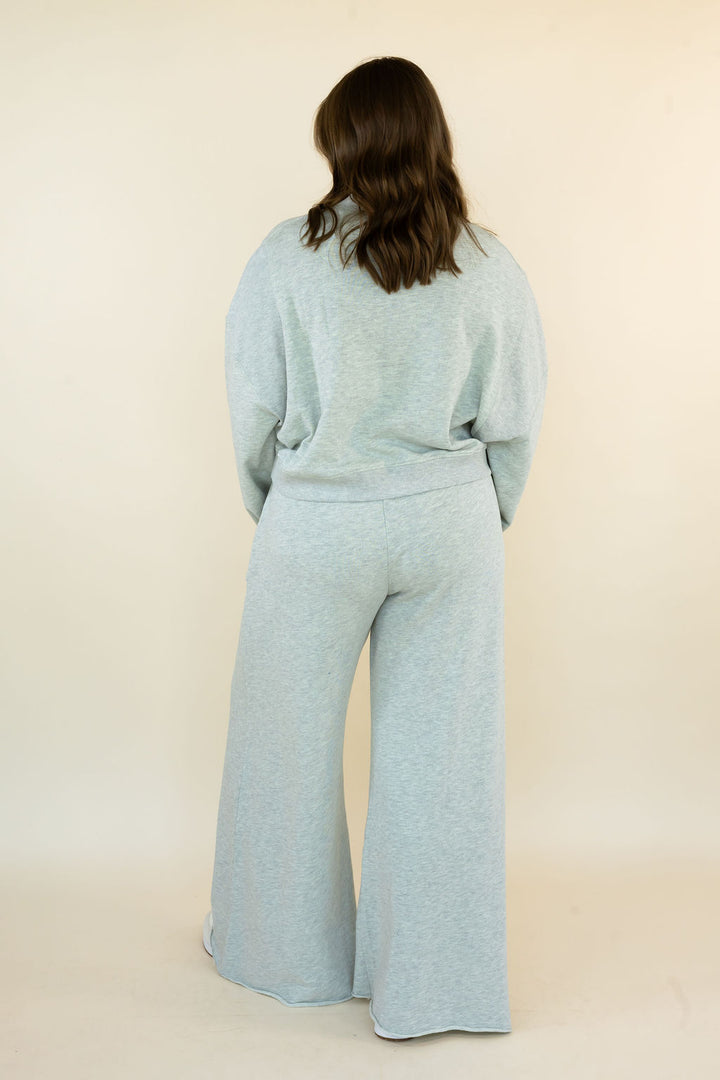 A woman viewed from behind wearing a light grey heathered lounge set consisting of a long-sleeve top and wide-leg pants, standing against a plain beige studio background.