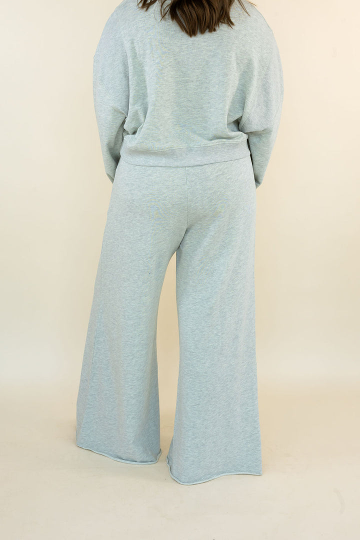 A person shown from the back wearing a light heathered grey matching lounge set with a long-sleeve top and wide-leg pants, against a plain cream studio background.