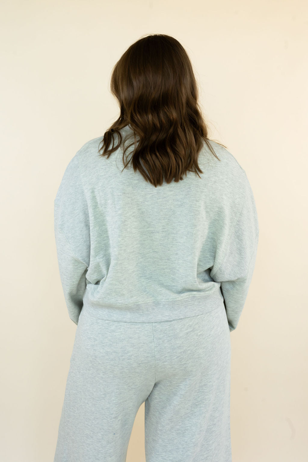 A person viewed from behind wearing a light grey heathered sweatshirt and matching sweatpants against a plain cream background.