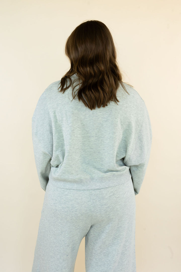 A person viewed from behind wearing a light grey heathered sweatshirt and matching sweatpants against a plain cream background.