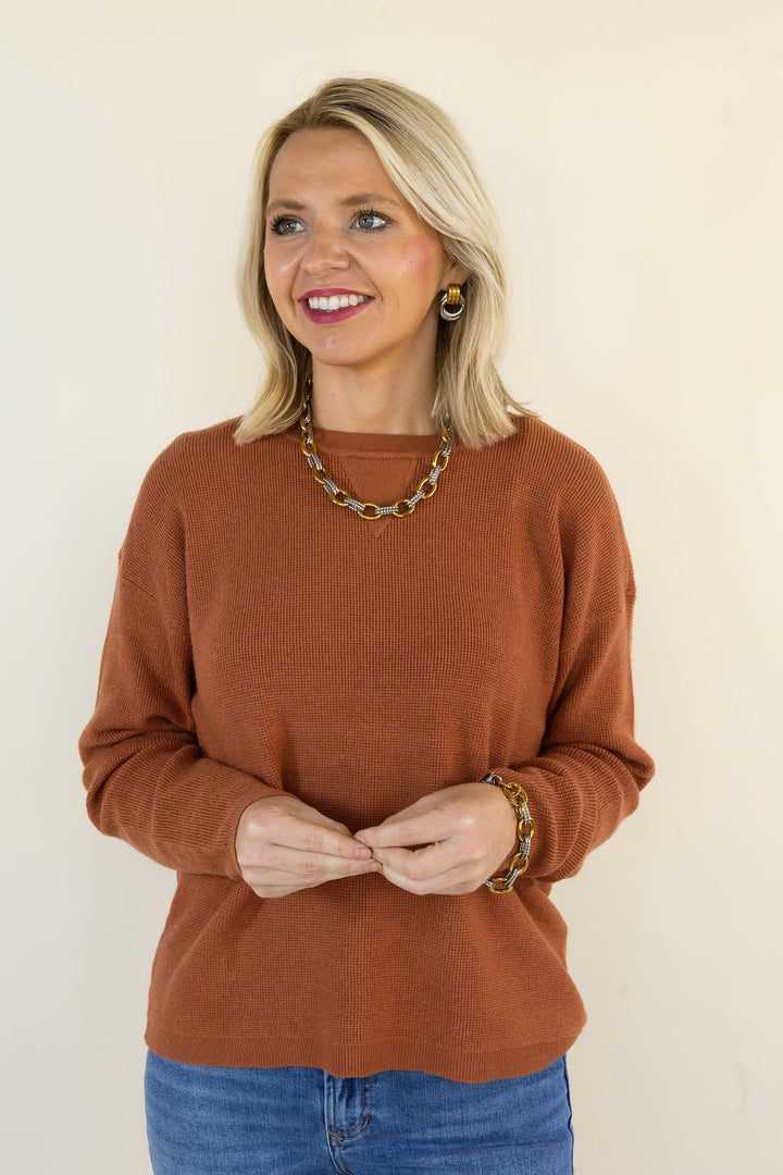 A woman with blonde hair wearing a rust-brown long-sleeve knit sweater, blue jeans, and a chunky gold chain necklace, standing against a plain beige background.