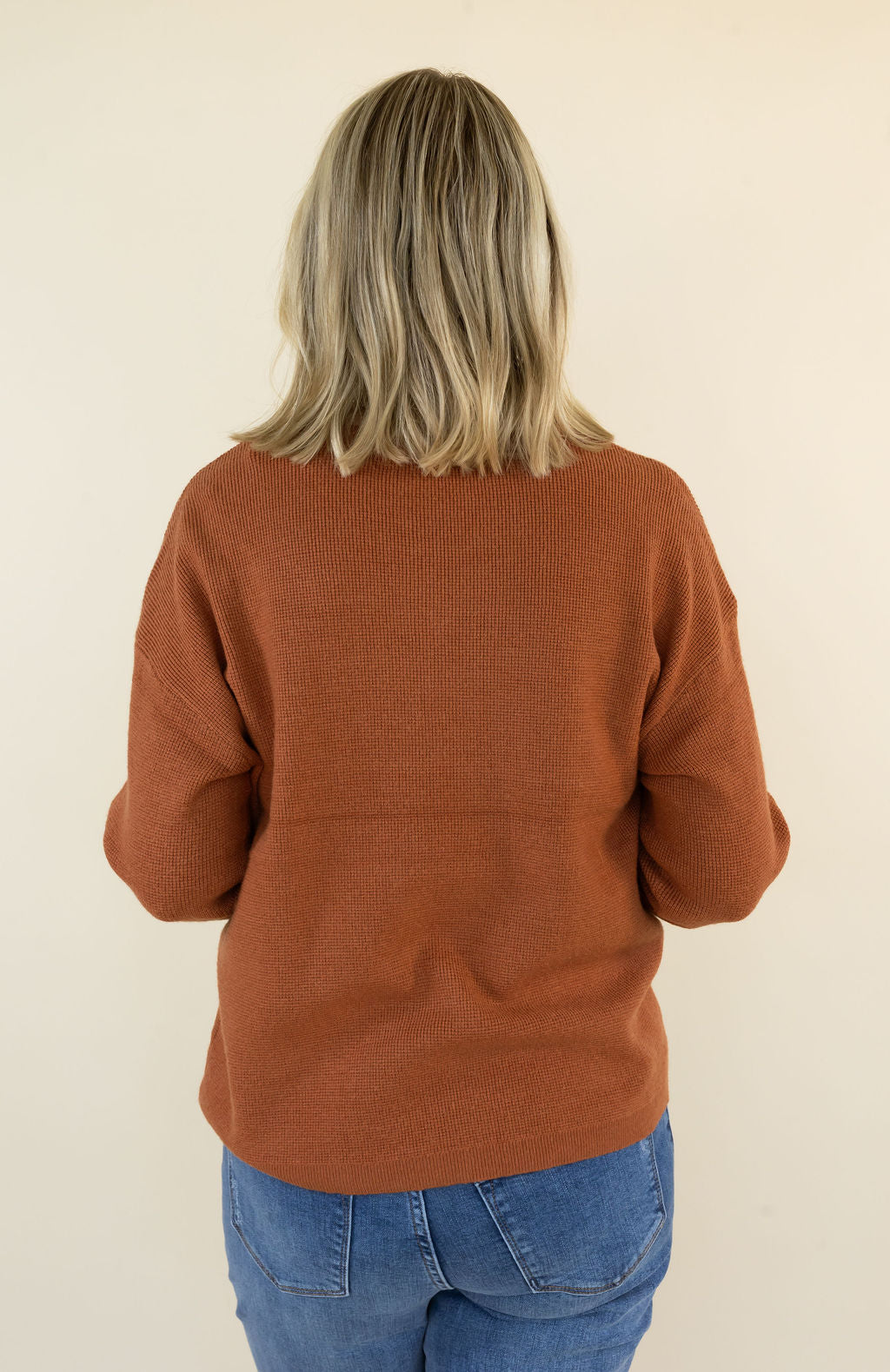 A woman viewed from behind wearing a rust-orange knit sweater and blue jeans, standing against a plain cream background.