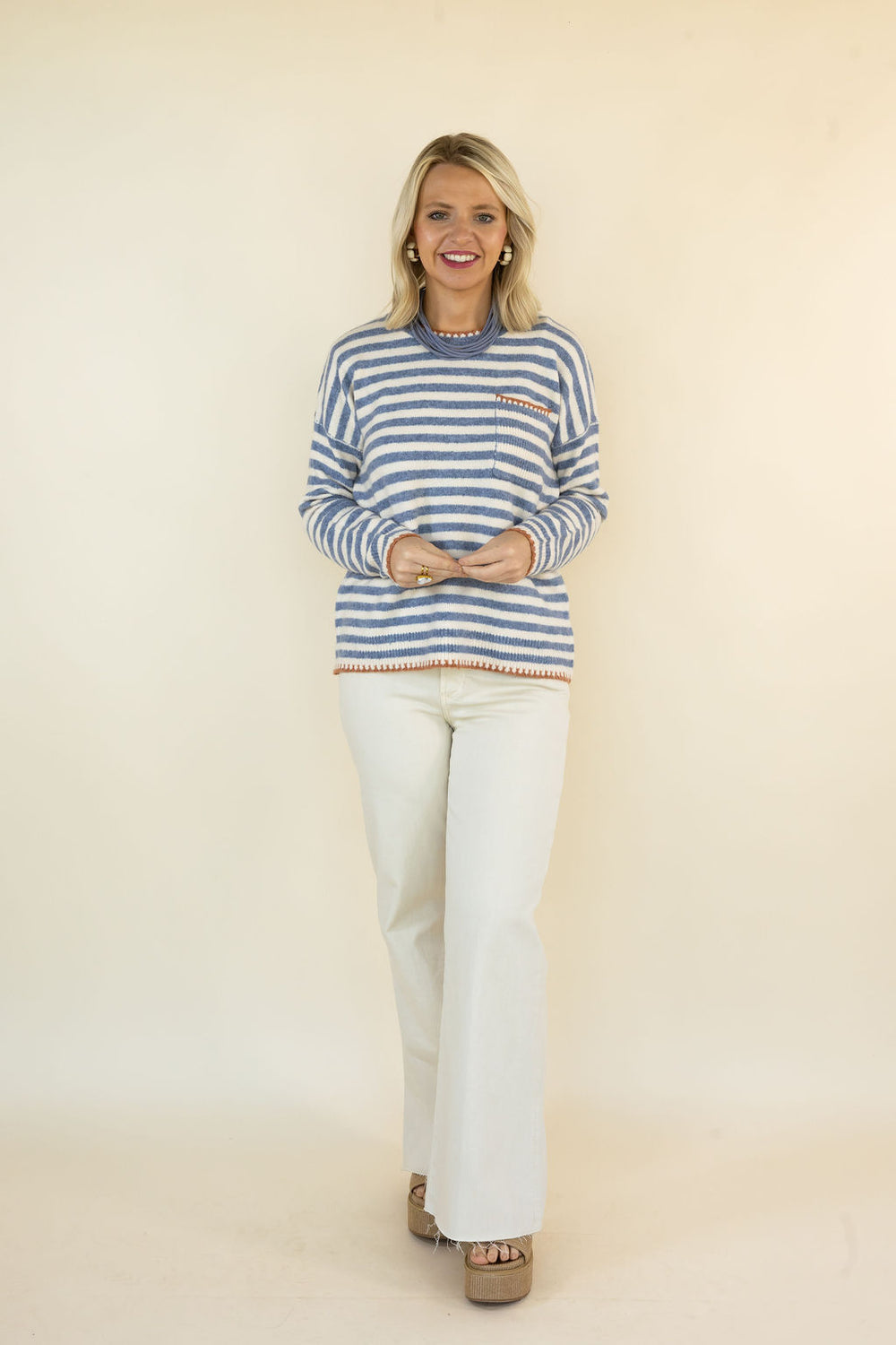 A woman modeling a blue and white striped knit sweater with orange trim, paired with white flared pants and brown platform sandals, standing against a solid beige studio background.