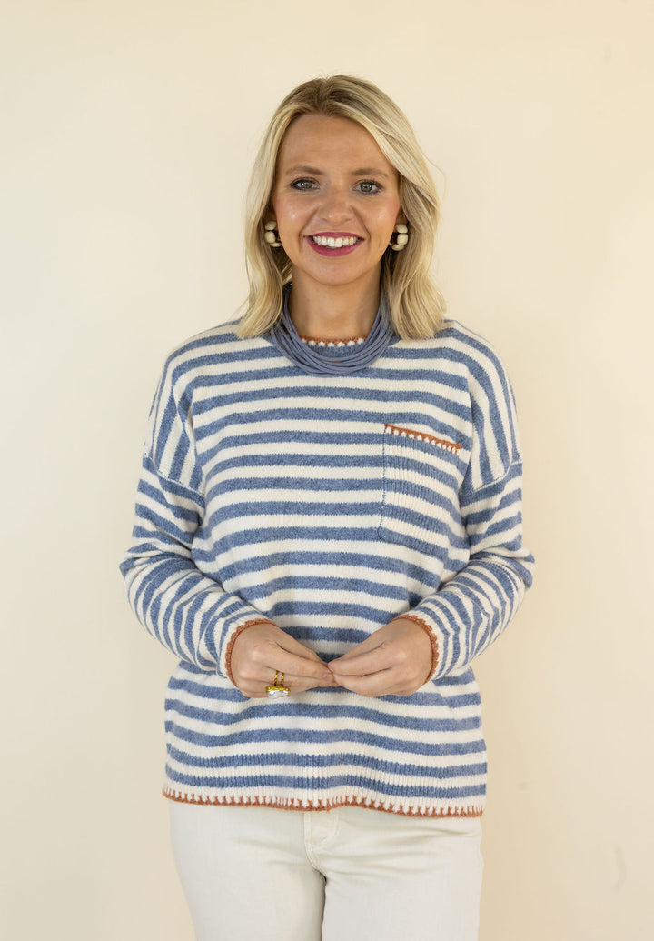 A smiling blonde woman wearing a blue and white striped knit sweater with a small chest pocket and rust-colored striped trim, paired with white pants, standing against a plain beige background.
