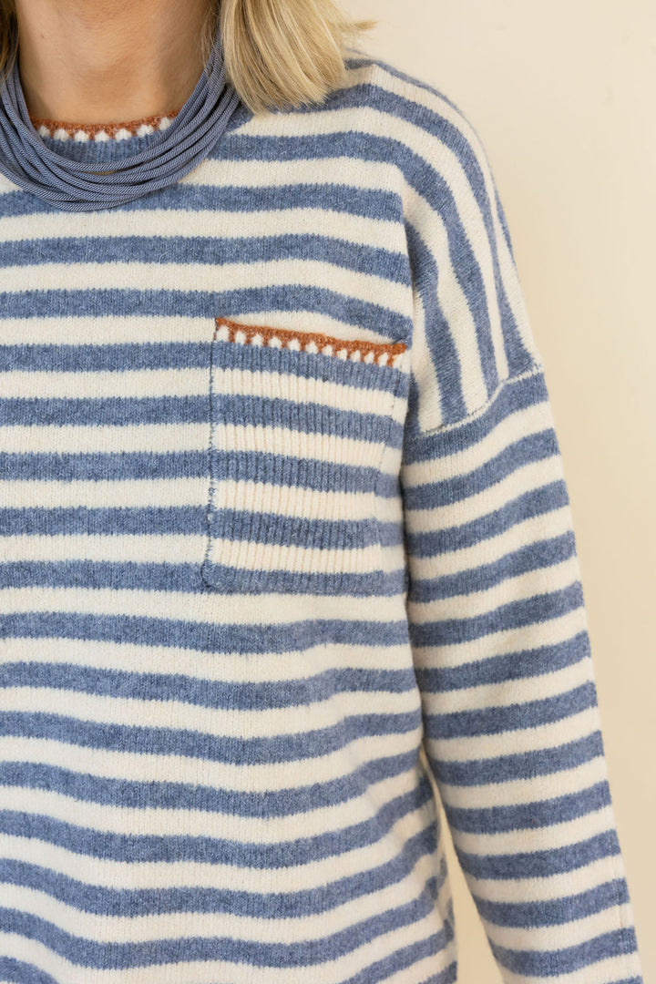 Close-up of a person wearing a blue and off-white striped knit sweater with a chest pocket and a small rust-colored decorative accent along the pocket edge.