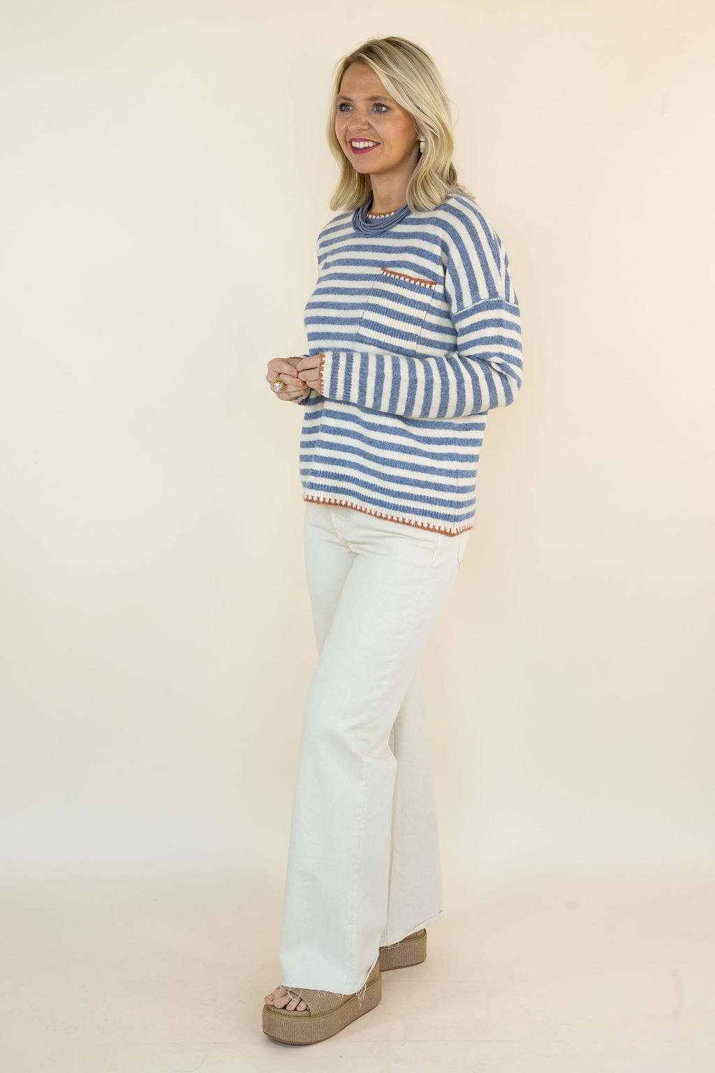 A woman modeling a blue and white striped knit sweater with a chest pocket, paired with white flared pants and brown platform sandals, against a plain cream studio background.