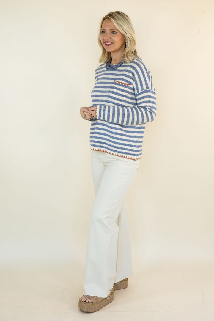 A woman modeling a blue and white striped knit sweater with a chest pocket, paired with white flared pants and brown platform sandals, against a plain cream studio background.