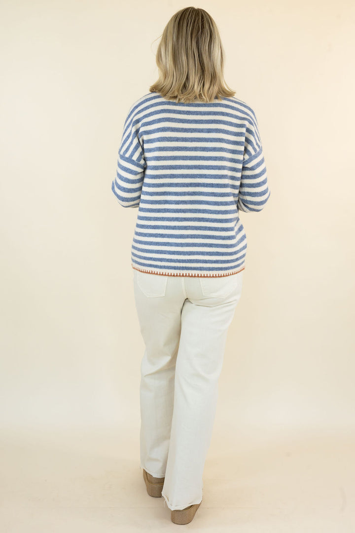 A woman viewed from behind wearing a blue and white striped knit sweater and off-white pants, standing against a plain beige studio background.