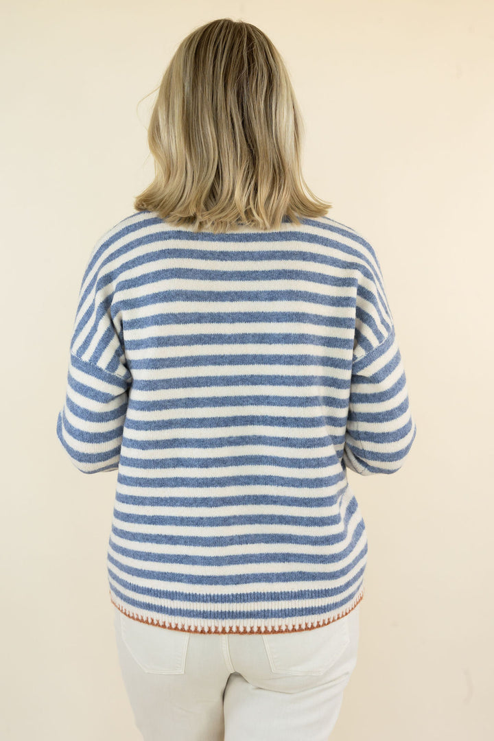 A woman viewed from behind wearing a blue and off-white striped knit sweater with a small rust-colored patterned hem, paired with off-white pants, against a plain beige background.
