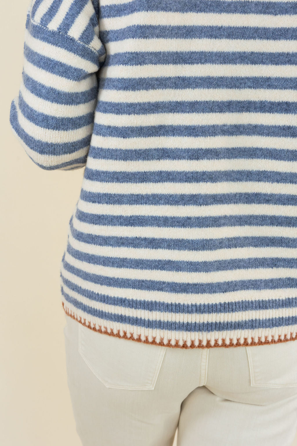 Close-up of a person wearing a blue and white striped knit sweater with a textured hem, paired with light beige pants, against a plain cream background.