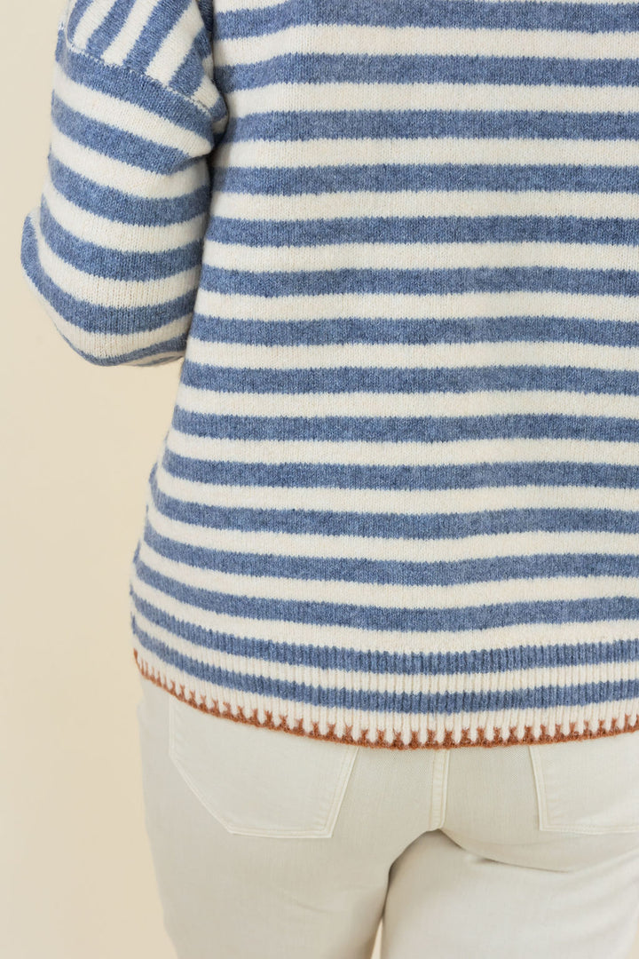Close-up of a person wearing a blue and white striped knit sweater with a textured hem, paired with light beige pants, against a plain cream background.