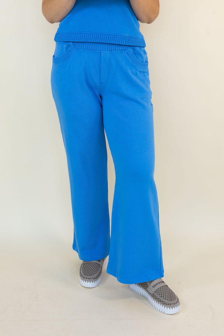 A person shown from the waist down wearing bright blue wide-leg pants and gray slip-on shoes against a plain light beige studio background.