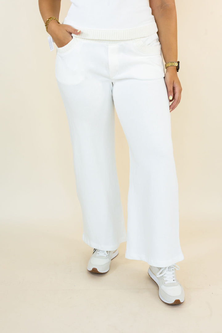 Ivory wide-leg cropped pants with a ribbed elastic waistband, modeled with white sneakers against a light beige studio background.