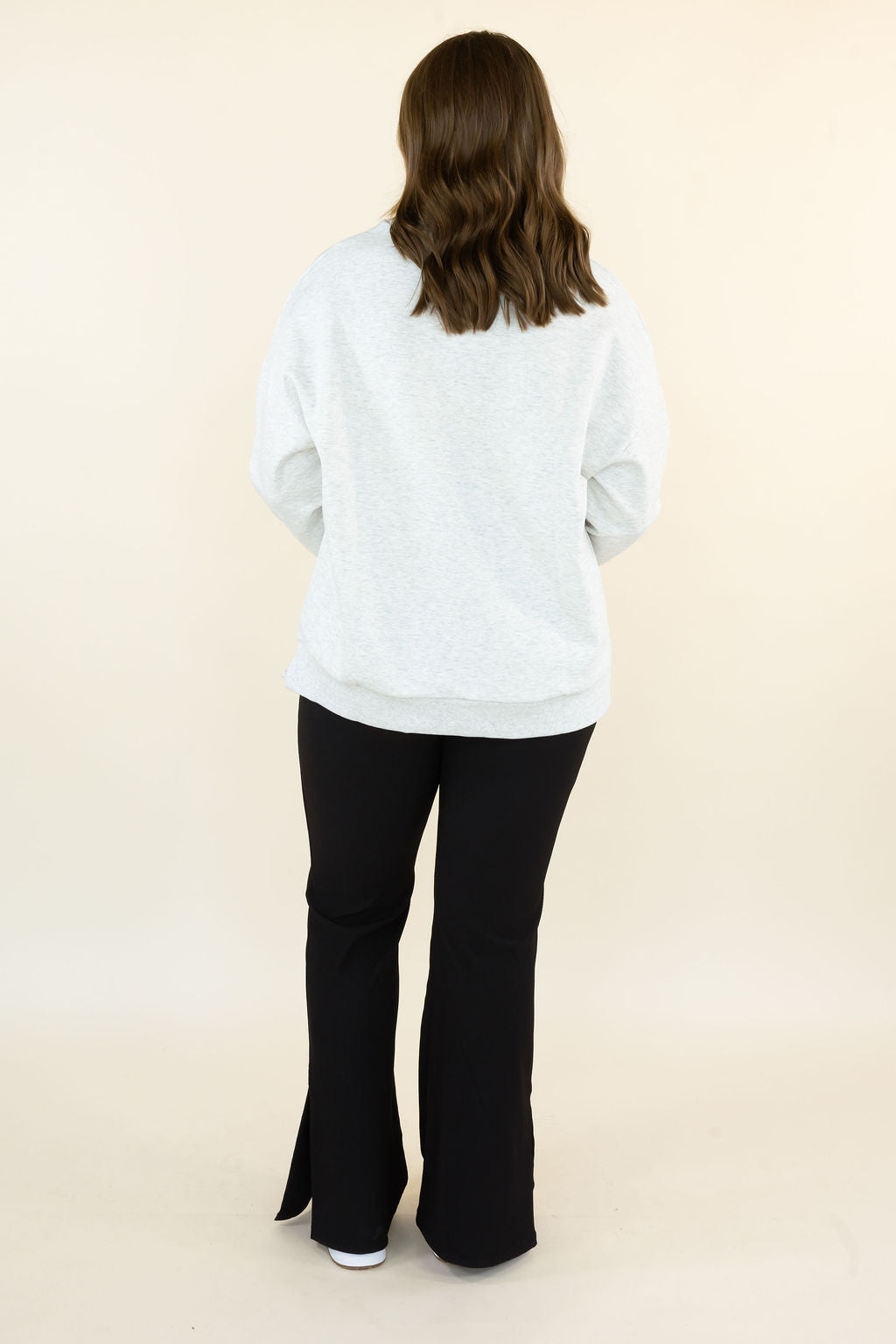 A woman viewed from behind wearing a light gray heathered sweatshirt and black flared pants, standing against a plain cream studio background.