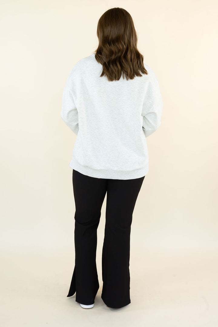 A woman viewed from behind wearing a light gray heathered sweatshirt and black flared pants, standing against a plain cream studio background.