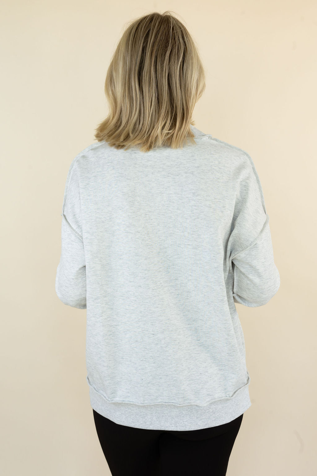 A woman viewed from behind wearing a light heather gray long-sleeve sweatshirt and black pants, standing against a plain beige background.