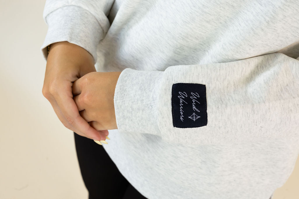 A close-up of a person wearing a light heather gray sweatshirt, focusing on the left sleeve which features a black square patch with white script text reading &