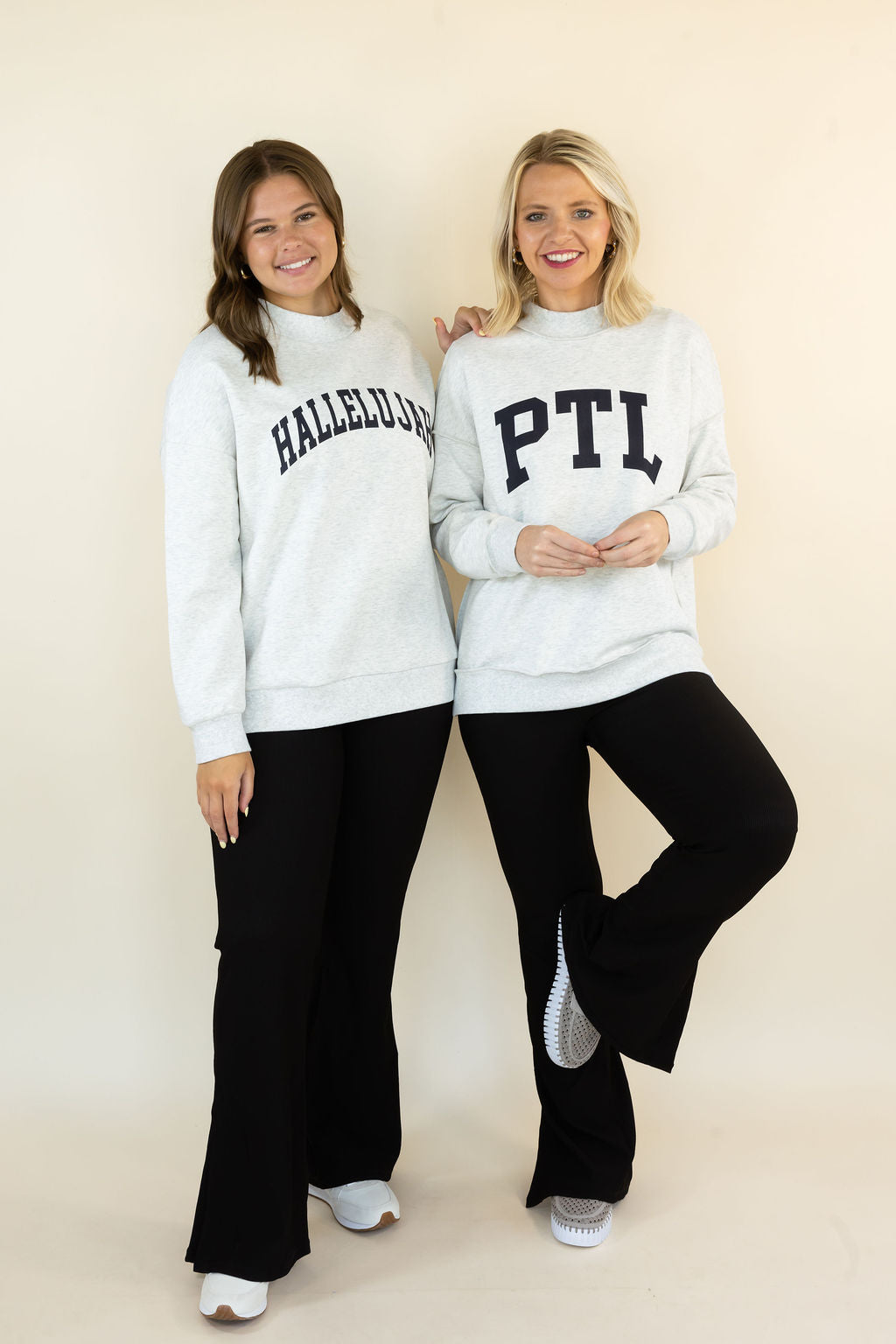 Two women modeling matching light gray sweatshirts with collegiate-style lettering, paired with black pants, standing against a plain beige studio background.