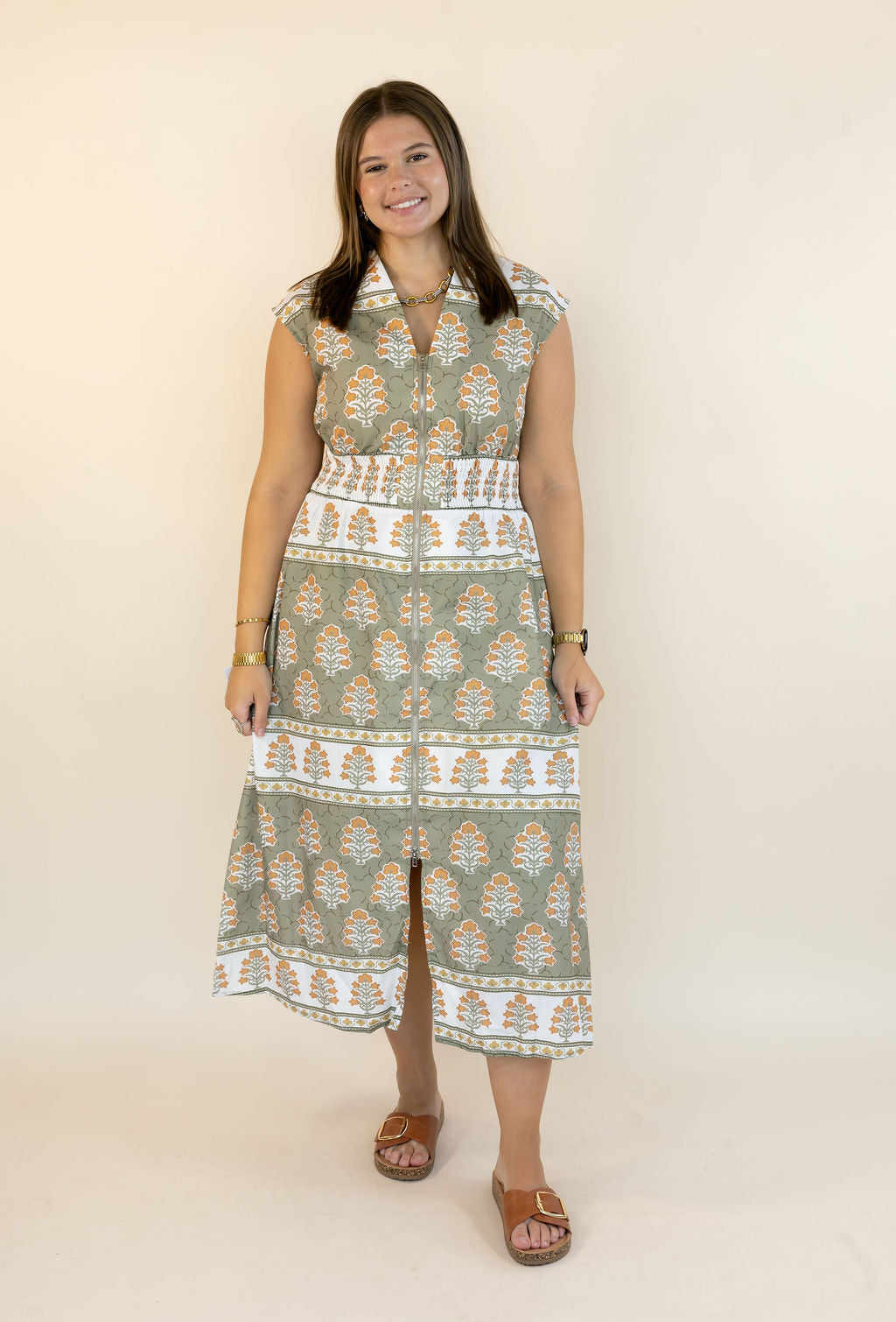 A woman modeling a sleeveless midi dress with a green, white, and orange geometric pattern, standing against a plain cream background.