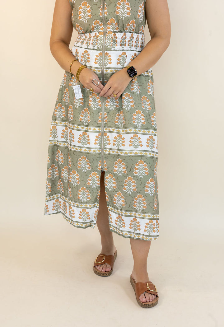 A sleeveless midi dress with a vertical front slit, featuring a sage green and white block print pattern with orange accents. The dress is modeled with brown slide sandals against a plain beige background.