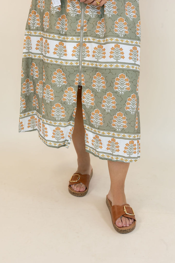 A person wearing a long-sleeve midi dress with a sage green, white, and orange botanical block print pattern, paired with brown slide sandals, shown from the waist down against a plain beige studio background.
