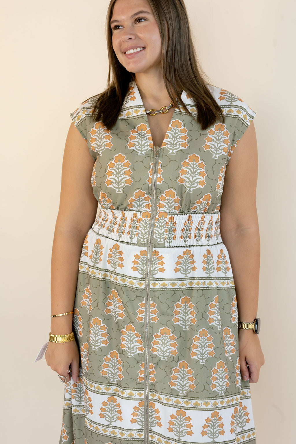 A woman wearing a sleeveless midi dress with a vertical front zipper, featuring a sage green, white, and orange block print pattern. She is standing against a plain beige wall.
