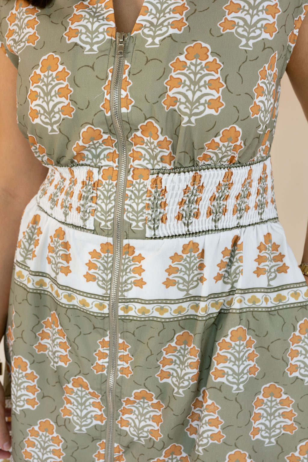 Close-up of a sleeveless dress with a front zipper, featuring an olive green, white, and orange floral block print pattern with a smocked waistband.