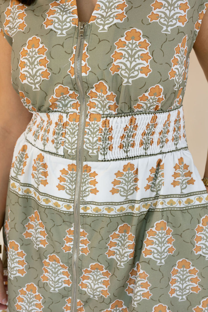 Close-up of a sleeveless dress with a front zipper, featuring an olive green, white, and orange floral block print pattern with a smocked waistband.