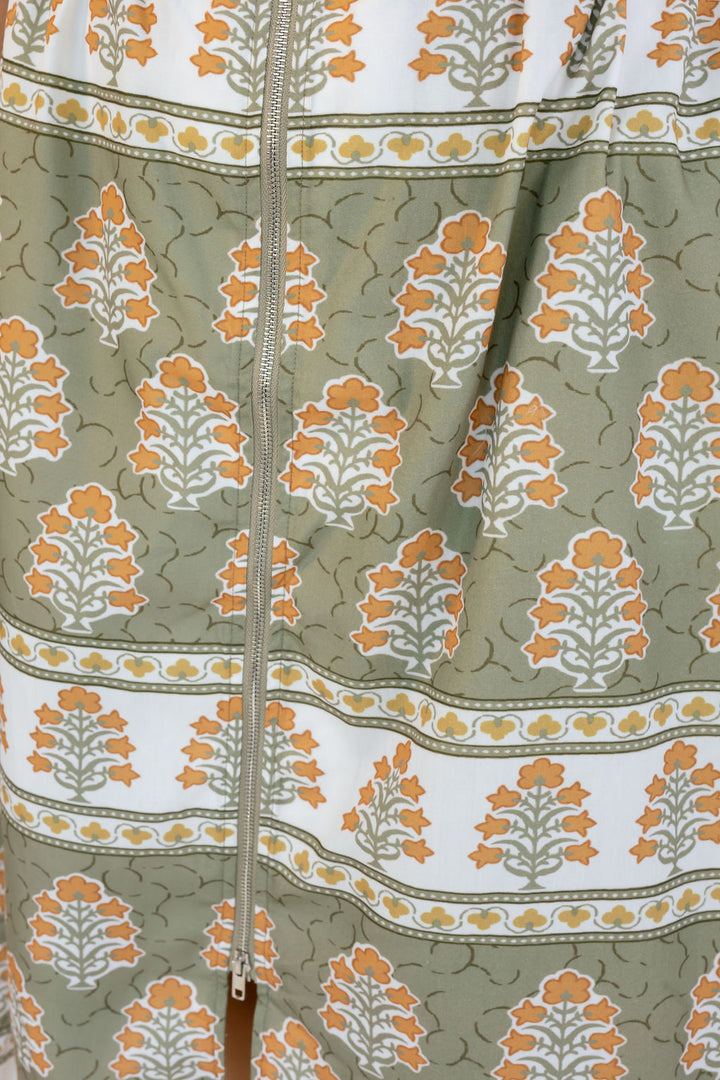 A close-up of a patterned garment featuring a sage green base with orange and white block-print style tree motifs and horizontal decorative borders, with a silver zipper running down the center.