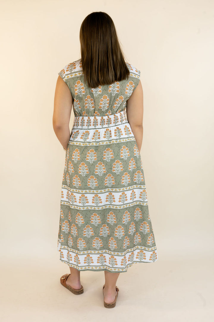 A woman viewed from behind wearing a sleeveless midi dress with a sage green, white, and orange block print pattern, paired with brown flat sandals, against a plain beige background.