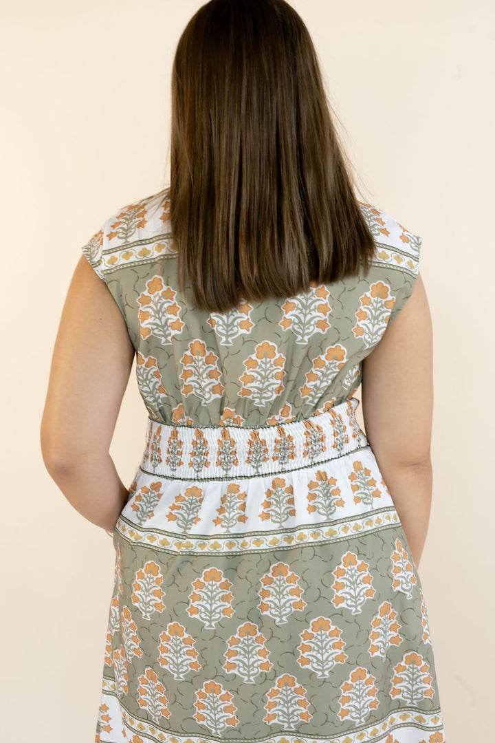 A woman viewed from behind wearing a sleeveless dress with a sage green, white, and orange block print pattern and a smocked waistband, set against a plain beige background.