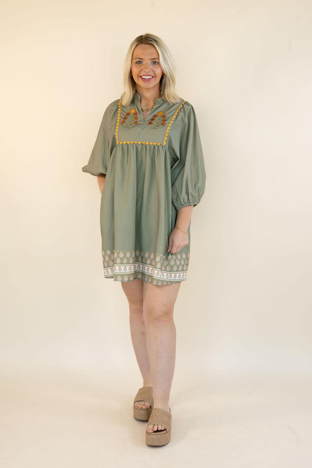 A woman modeling a sage green short-sleeve tunic dress with yellow embroidered details on the chest and a patterned hem, standing against a plain cream background.