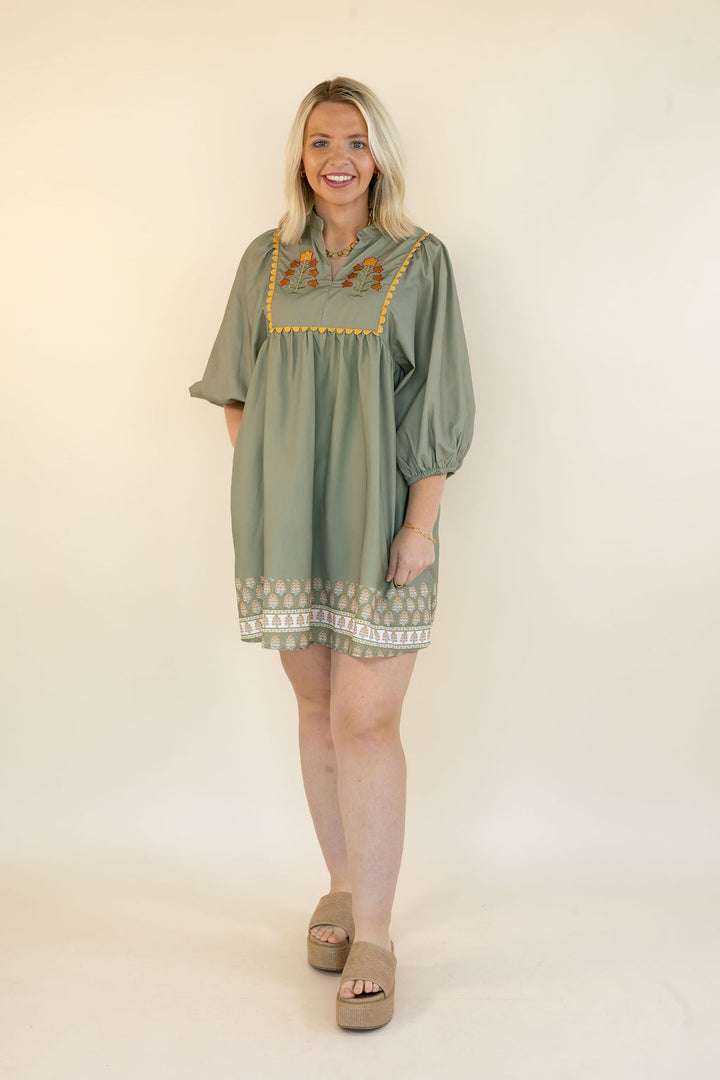 A woman modeling a sage green short-sleeve tunic dress with yellow embroidered details on the chest and a patterned hem, standing against a plain cream background.