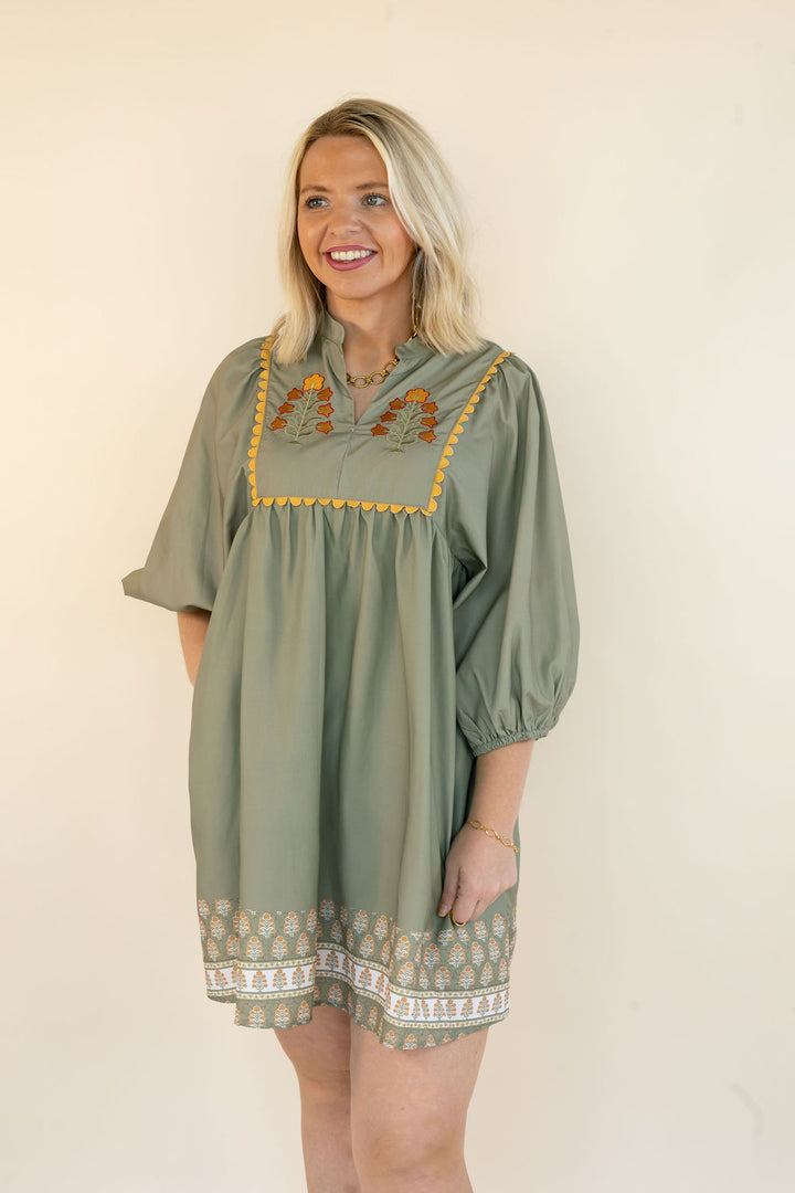 A woman modeling a loose sage green tunic dress with yellow embroidered details on the chest and a patterned hem, set against a plain cream background.