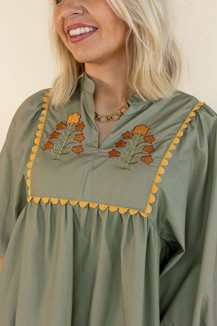 A close-up of a woman wearing a sage green blouse with orange and yellow floral embroidery and yellow scalloped trim around the neckline.