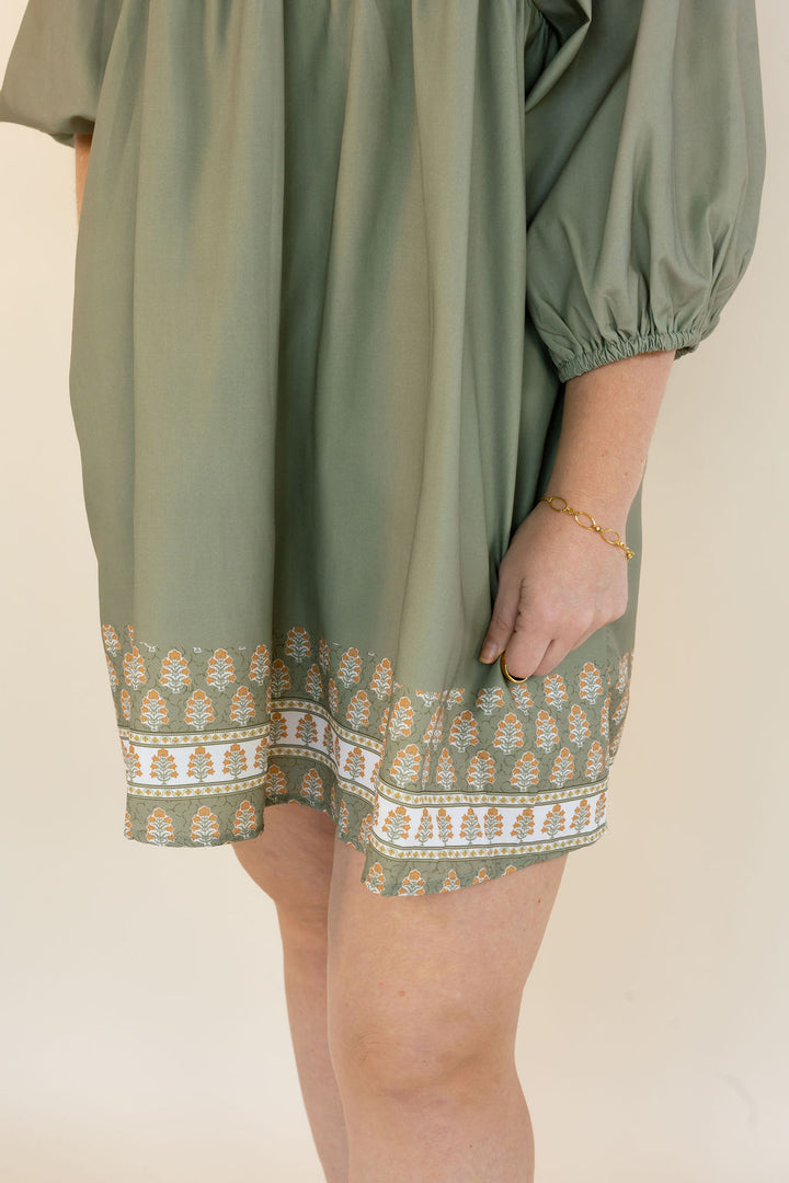 An olive green dress with a patterned hem and a gold bracelet on a model&
