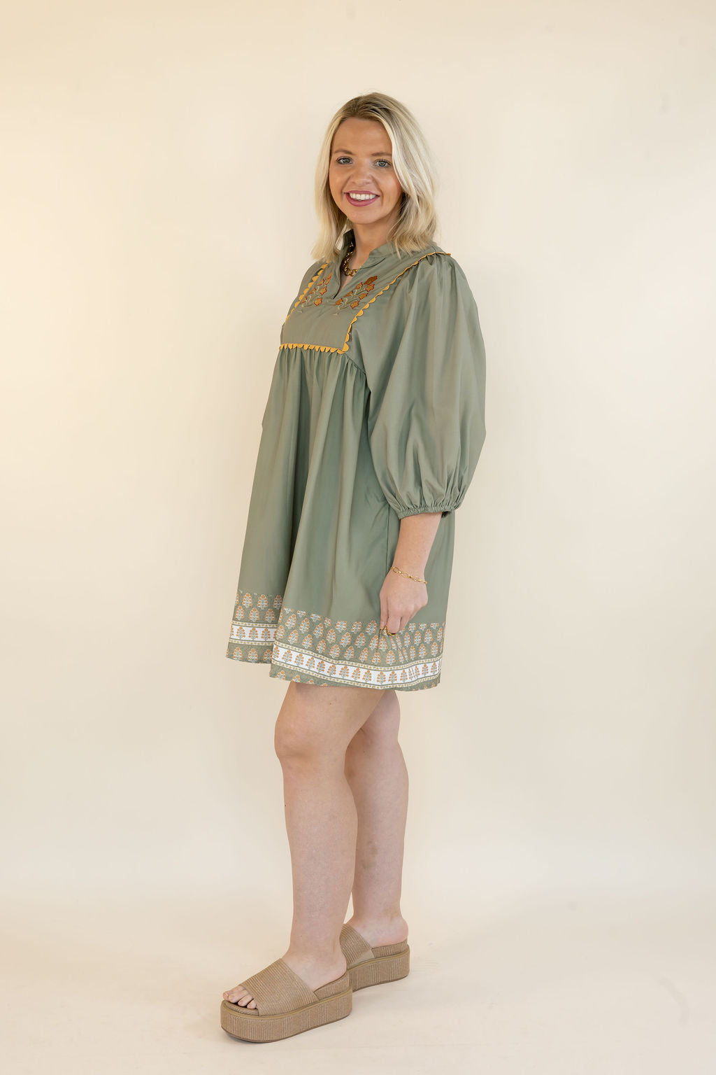 A woman modeling a sage green short dress with gold embroidered detailing on the chest and a white patterned hem, standing against a plain cream background.